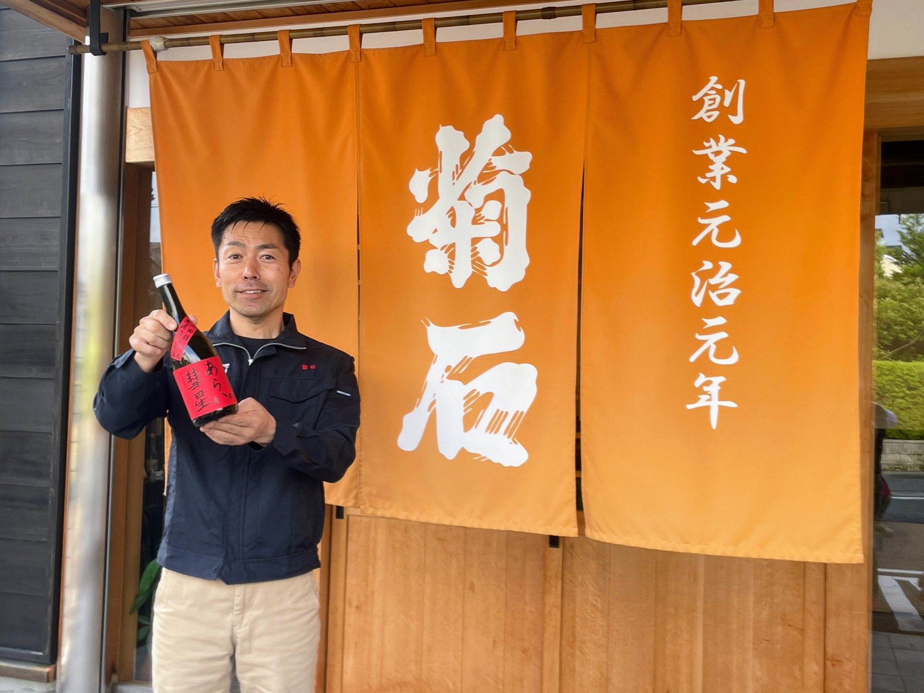 オレンジ色の暖簾の前で日本酒を持って立つ男性