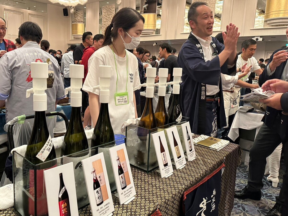 テーブルの上に日本酒が並んでいる後ろで説明する男性と女性